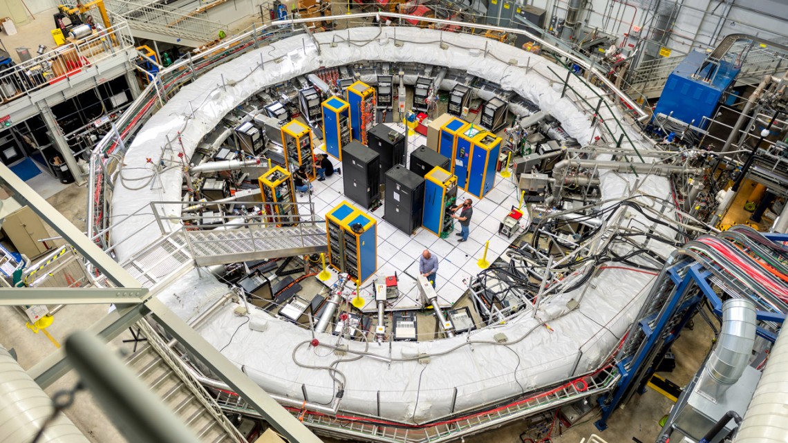 The muon g-2 ring sits in its detector hall amidst electronics racks, the muon beamline and other equipment at the Fermi National Accelerator Laboratory.